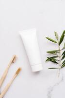 Top view white plastic tube without label, bamboo toothbrushes and olive branch on white background photo