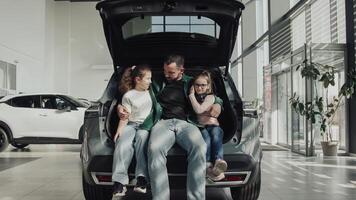 A happy father with his two daughters sits in the trunk of the car he just bought. The happy family is enjoying the purchase. The end of a test drive or purchase of a new, modern car. photo