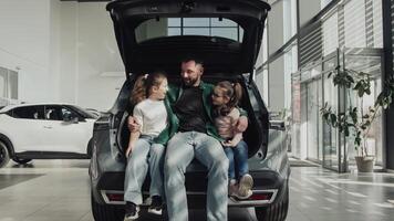 A happy father with his two daughters sits in the trunk of the car he just bought. The happy family is enjoying the purchase. The end of a test drive or purchase of a new, modern car photo