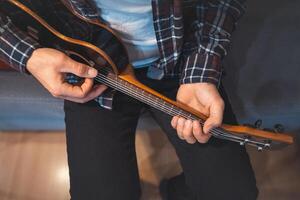 A man in a denim jacket sits on a blue sofa and strums a small wooden ukulele under warm indoor lighting. photo