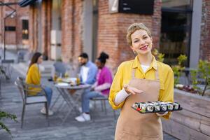 Smiling waitress presents a plate of sushi in an outdoor restaurant setting, with customers in the background. photo