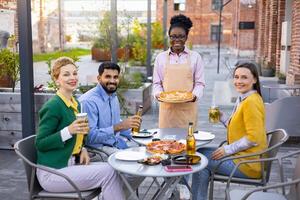 A diverse group enjoys pizza and drinks at an outdoor restaurant, served by a smiling waitress. The scene captures the joy of social gathering. photo