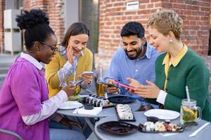 A diverse group of friends are gathered at an outdoor cafe, enjoying food and using their smartphones. photo