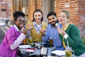 A diverse group of friends enjoys a sushi meal outdoors, smiling and using chopsticks at a table. photo
