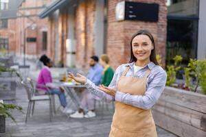 Smiling waitress welcomes customers to an outdoor restaurant, gesturing towards the seating area with other patrons. photo