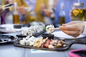 Close-up of a hand using chopsticks to eat sushi from a table set with other food and drinks. Friends enjoy the meal. photo