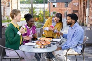 A diverse group of friends enjoys pizza and drinks at an outdoor restaurant, capturing a moment together with a photo. photo