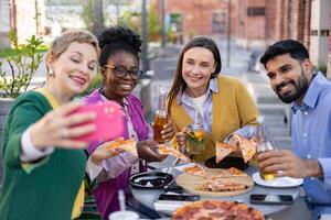 A diverse group of friends enjoys pizza, drinks, and taking selfies outdoors on a sunny day. photo