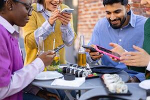 A diverse group of friends enjoy sushi and drinks while looking at their phones, sharing moments together at an outdoor table. photo