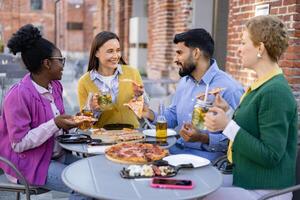 A diverse group of friends enjoys pizza and drinks at an outdoor restaurant, laughing and conversing around a table. photo