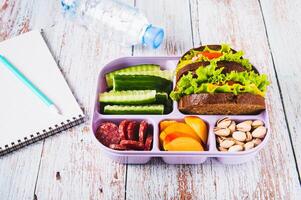 Lunchbox with sandwich, chorizo, cucumber and peach, bottle of water on table photo