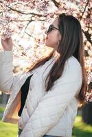 Woman enjoying cherry blossoms in springtime park while wearing sunglasses and a light jacket photo