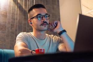Focused young man wearing glasses and wireless earbuds, watching a webinar, having a call with a client or a business conference meeting on a laptop in a modern interior with ambient light. photo