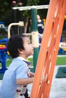 Little boy playing happily on the playground in the summer sun. Asian kid climb the orange play equipment for climbing, play and development of small and large muscles, decision making and courage. photo