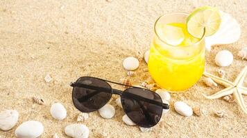 Summer concept with cold refreshing orange or lemon drink, sunglasses, starfish, and sea shells isolated on beach sand background photo