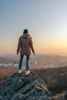 A girl in a jacket and a hat stands on a rock and looks at the city from a height during sunset. photo