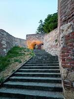 Stone steps lead to an illuminated archway in an old fortress. The path is flanked by historic brick and stone walls, leading the viewer into a serene twilight setting photo
