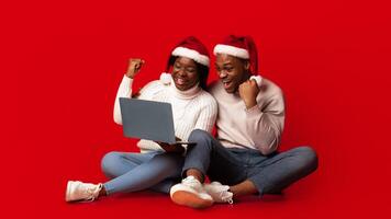 Christmas Offer. Excited black couple in santa hats celebrating success with laptop over orange background, cheerful african spouses sitting with computer and raising fists with joy, copy space photo