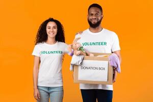 Couple Of Volunteers Posing With Donation Box Peeking Clothes For Charity Over Yellow Background, Smiling To Camera. Studio Shot. Volunteering Work, Contribution Concept photo