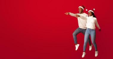 Promo Offer. Cheerful Black Couple In Santa Hats Pointing At Copy Space, Demonstrating Free Place On Orange Background For Your Christmas Holiday Advertisement, Jumping Together And Having Fun photo