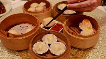 A hand uses chopsticks to pick dumplings from bamboo steamers on a restaurant table with sauces video