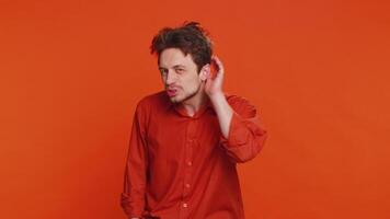 I cant hear you. What. Caucasian man trying hear you looking confused and frowning keeping arm near ear for louder voice asking to repeat, to hear information, deafness. Young guy on orange background video
