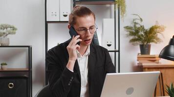 Happy Caucasian stylish young businessman communicating with customer by phone smartphone, working on laptop netbook at home office desk. Guy in suit represents concept of distance job on Internet. video