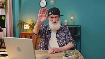 Hello. Happy stylish senior man employee waves hand hi gesture greeting welcomes someone, online webinar, remote distance conversation. Freelancer grandpa with laptop netbook at desk in home office video