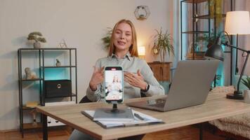 Smiling middle-aged business woman sitting at table before smartphone fixed on tripod record motivation training webinar on web camera. blogger influencer broadcast live speech using cell phone. video