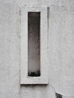 Close up of rectangular wall vent embedded in concrete wall, showing signs of weathering and dirt accumulation inside vent opening, with visible cracks and rough texture on wall surface photo