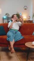 Happy young Caucasian woman making online call on smartphone talking waving hand hello. Communication, meeting. Smiling girl making cellphone conversation sitting on sofa couch at home. Vertical video