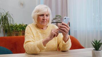 Senior woman use smartphone typing browsing, loses becoming surprised sudden lottery results, bad news, fortune loss, fail. Elderly mature grandmother at home living room sits on couch at table desk video
