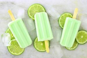 Healthy lime yogurt popsicles with fresh lime slices scattered on a white marble background photo