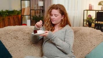 Pregnant young woman sits on home sofa, holding a slice of cake, smiling with satisfied expression, enjoying sweet taste. Girl embraces small pleasures, finding joy and comfort in simple happy moments video