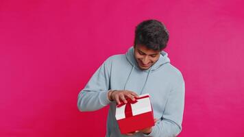 Excited Indian man receiving a festive gift with a red bow opening the box with joy celebrating a surprise. Happy Arabian guy enjoying dream present smiling with excitement isolated on pink background video