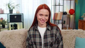 Young woman at home winking at camera with flirty smile showing playful confident mood and charm. Caucasian redhead girl on sofa enjoying fun moment, expressing emotion and personal attractive energy video