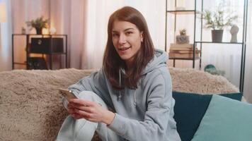 Young woman using smartphone typing browsing making recommendation smiling at camera. Pretty happy adult girl in casual clothes showing like thumbs up at home in living room apartment sitting on sofa. video