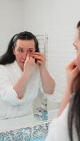 Middle-aged mature woman comfortably standing at mirror applying eye patches, gently smoothing them with fingers. Lady smiling softly at reflection, enjoying relaxing moment of personal skincare video