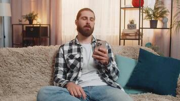 Young man sits at home with smartphone, feeling sudden disappointment from mobile game defeat. Guy on sofa exhales heavily, revealing gloom, frustration, bad news, fortune loss, fail negative result video