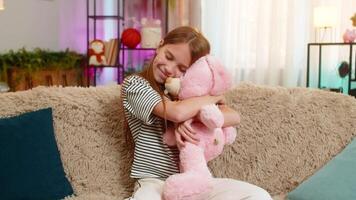 Young child girl lovingly hugs a soft bunny toy while sitting at home on the sofa, smiling with joy tenderness. School kid shows affection, comfort, emotional warmth during playful quiet time indoors video