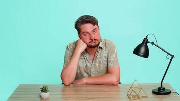 Middle-aged man sits at table with blank stare, head supported by hand, showing emotional burnout, tiredness, complete apathy. Young adult guy isolated on blue background expressing exhaustion fatigue video