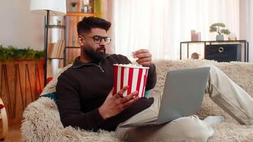 Indian man at home watches laptop while eating popcorn, laughing during movie scene with joyful face. Hispanic guy on sofa feels happy and relaxed, enjoying peaceful moment of fun and digital comfort. video