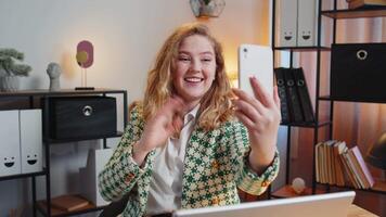 Hello. Happy businesswoman waves hand hi gesture greeting welcomes someone on smartphone, online selfie webinar, remote distance conversation. Freelance sitting at desk using laptop in home office video