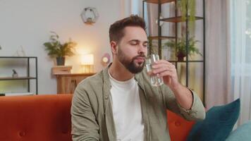 Satisfied young Caucasian brunette man sitting at home on sofa leading healthy lifestyle, drinking a glass of pure water, smiling. Drink daily norm of water. Guy smiling gestures at camera in room. video