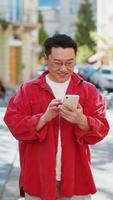 Asian middle-aged man using smartphone typing text messages in social media application online, surfing internet, relaxing, taking a break outdoors. Chinese guy walking on urban city street. Vertical video