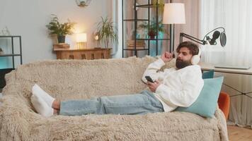 Happy overjoyed young man in wireless headphones with smartphone relaxing sitting on couch at home apartment choosing, listening favorite energetic disco dancing music. Weekend leisure activities. video