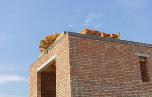 A brick structure stands incomplete with wooden beams positioned on its roof under a bright blue sky, showcasing ongoing construction work photo