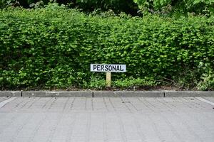 In front of a green hedge, there is a white sign with black lettering saying Personal in a parking lot photo