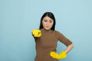 Frustrated female cleaner in yellow rubber gloves pointing finger directly at camera, expressing anger and emotional pressure. Upset housekeeper confronting viewer over blue background. photo