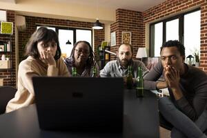 Four diverse young adults staring at laptop screen in surprise while watching suspenseful movie. Group friends with green beer bottles on table, suggesting casual indoor hangout in cozy apartment. photo